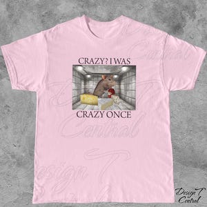 Puede incluir: Camiseta rosa claro con un gr&aacute;fico que presenta una rata de dibujos animados en una habitaci&oacute;n con queso. El texto de la camiseta dice "CRAZY? I WAS CRAZY ONCE". La camiseta es de cuello redondo.