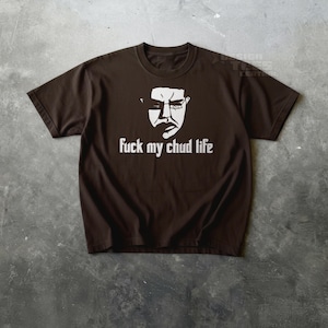 May include: A brown t-shirt featuring a white graphic of a face with glasses and the phrase "fuck my chud life" in white block letters. The shirt is made of a soft material and has short sleeves.