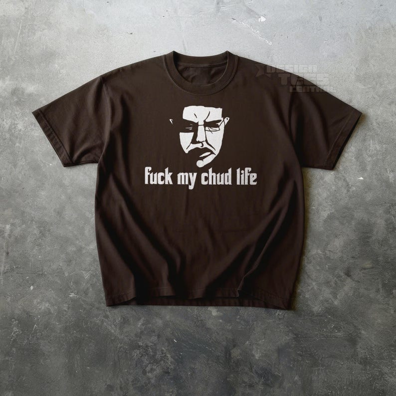 May include: A brown t-shirt featuring a white graphic of a face with glasses and the phrase "fuck my chud life" in white block letters. The shirt is made of a soft material and has short sleeves.