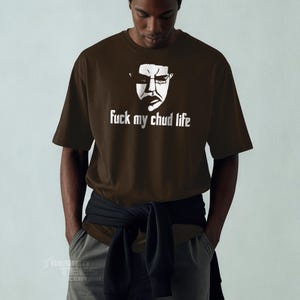 May include: A brown t-shirt with a white graphic of a face wearing glasses and the text "fuck my chud life". The shirt is short-sleeved and worn by a person with their hands in their pockets.
