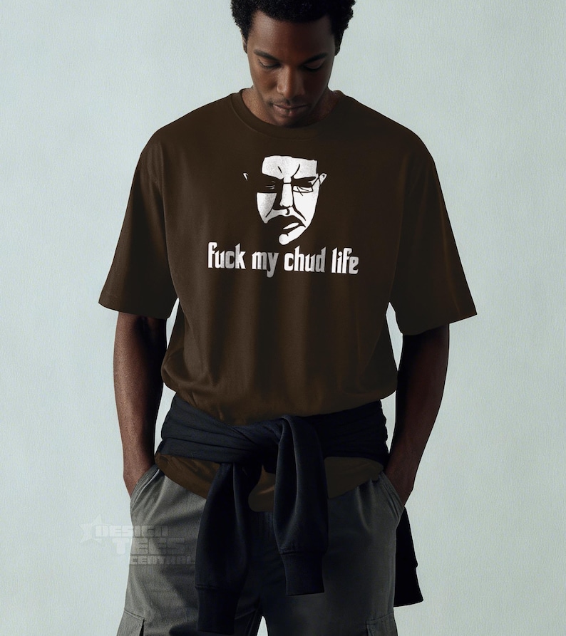 May include: A brown t-shirt with a white graphic of a face wearing glasses and the text "fuck my chud life". The shirt is short-sleeved and worn by a person with their hands in their pockets.