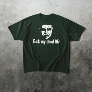 May include: Dark green t-shirt with a white graphic of a face and the text "fuck my chud life". The shirt is made of a soft material and has short sleeves.