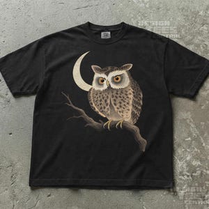 Może przedstawiać: Czarny t-shirt z krótkim rękawem z nadrukiem graficznym przedstawiającym brązowo-szarą sowę siedzącą na gałęzi drzewa na tle kremowego półksiężyca. Sowa ma duże pomarańczowe oczy i szczegółowe wzory piór na całym ciele.