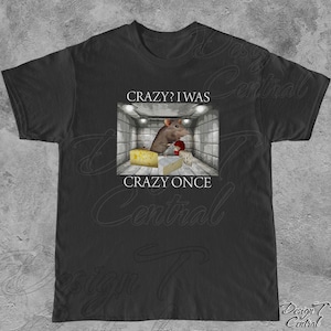 Puede incluir: Camiseta negra con un gr&aacute;fico de una rata en una habitaci&oacute;n con queso, con el texto "Crazy? I was crazy once". El dise&ntilde;o incluye una rata sosteniendo un objeto rojo y varios tipos de queso.