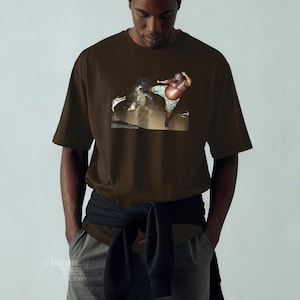 May include: Brown t-shirt featuring a graphic of a person in camouflage shorts kicking in water. The t-shirt is a solid brown color. The person in the image is dark-skinned.