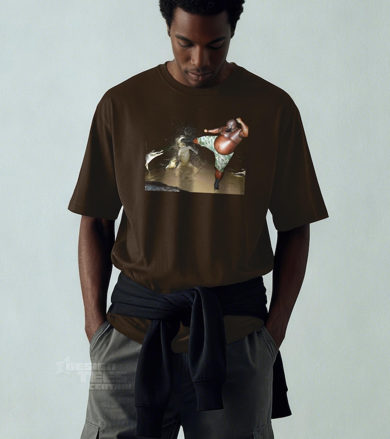 May include: Brown t-shirt featuring a graphic of a person in camouflage shorts kicking in water. The t-shirt is a solid brown color. The person in the image is dark-skinned.