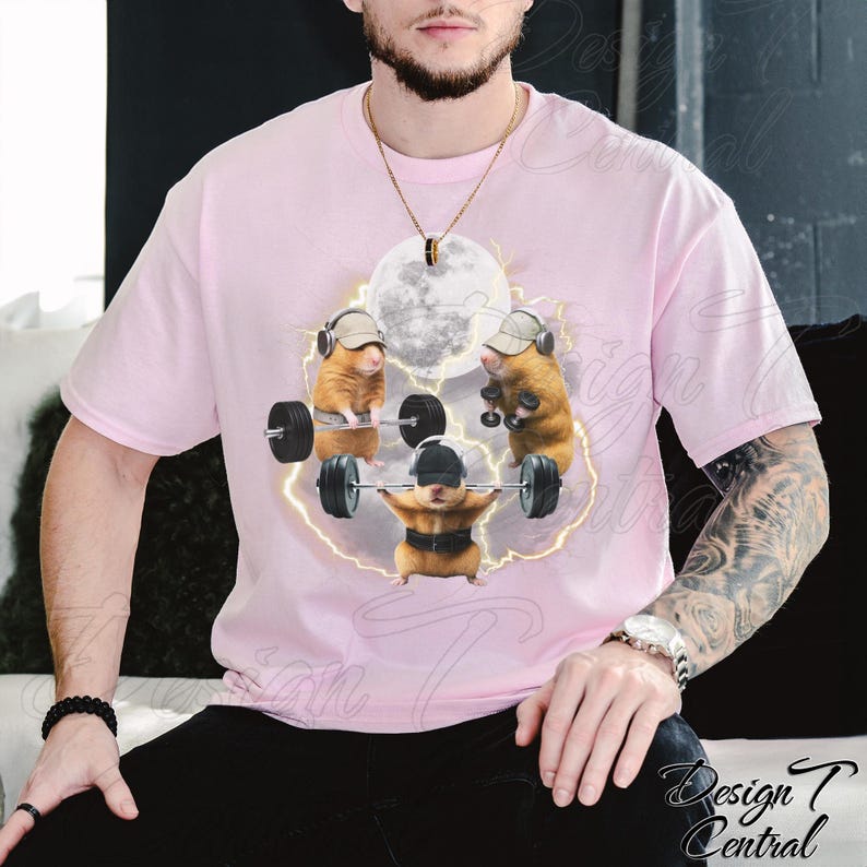 May include: A light pink t-shirt featuring a graphic of three hamsters lifting weights under a full moon. The hamsters are wearing hats and headphones. The design is surrounded by a lightning bolt effect.