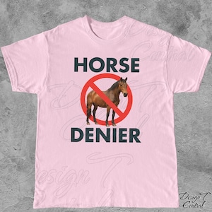 Peut inclure: T-shirt rose clair avec l'inscription "HORSE DENIER" en vert fonc&eacute;. Un cheval brun est &agrave; l'int&eacute;rieur d'un cercle rouge barr&eacute; d'une ligne rouge. Le t-shirt est &agrave; col rond.