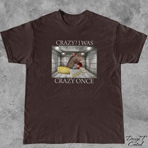 Puede incluir: Camiseta marr&oacute;n con un dise&ntilde;o humor&iacute;stico. El dise&ntilde;o muestra una rata de dibujos animados en una habitaci&oacute;n con queso y guantes de boxeo, con el texto "CRAZY? I WAS CRAZY ONCE".