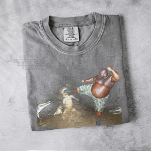 May include: Gray Comfort Colors t-shirt featuring a graphic of a person kicking a crocodile. The person is wearing camouflage shorts. The shirt has a tag that reads "Comfort Colors" and the size "S - P - CH".