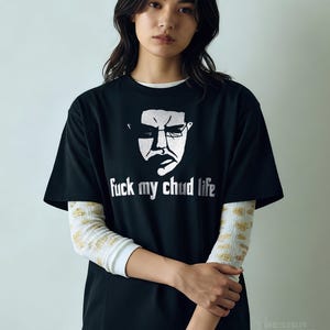 May include: Black t-shirt with a white graphic of a face and the text "fuck my chud life". The shirt is worn over a white long-sleeved shirt with a gold pattern. The design is from Design Tees Central.