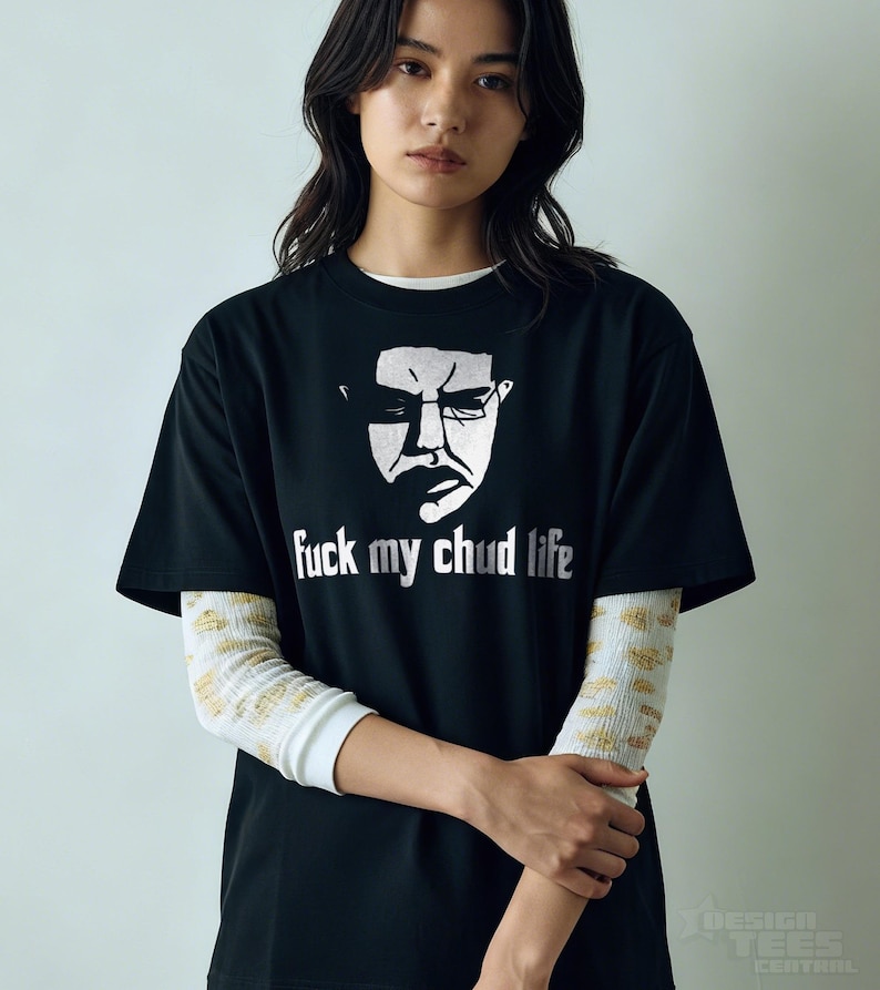 May include: Black t-shirt with a white graphic of a face and the text "fuck my chud life". The shirt is worn over a white long-sleeved shirt with a gold pattern. The design is from Design Tees Central.