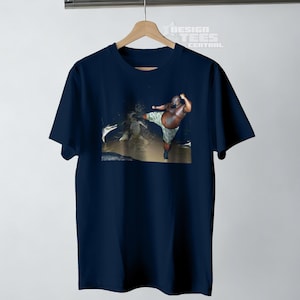May include: Navy blue t-shirt featuring a graphic of a person kicking. The person is wearing camouflage shorts. The shirt is hanging on a wooden hanger. The text "DESIGN TEES CENTRAL" is visible.