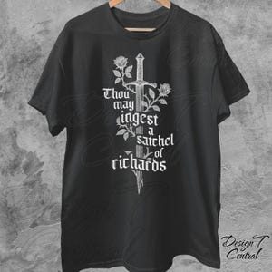以下が含まれることがあります： 黒いTシャツ。白いグラフィックは剣、バラ、そして「Thou may ingest a satchel of richards」というテキストが特徴です。シャツは木製のハンガーに展示されています。