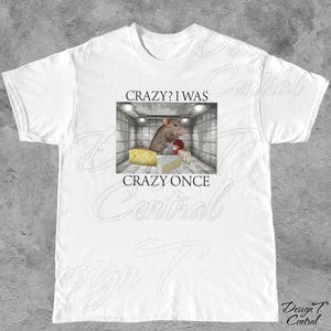 Puede incluir: Camiseta blanca con un gr&aacute;fico que presenta una rata de dibujos animados en un entorno de instituci&oacute;n mental, con queso y el texto "CRAZY? I WAS CRAZY ONCE". El dise&ntilde;o est&aacute; centrado en la camiseta.