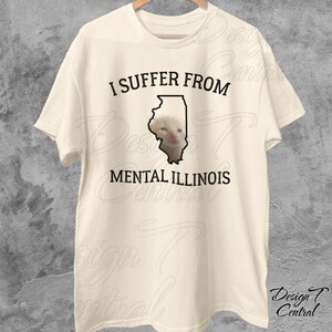 May include: A beige t-shirt with black text that reads "I suffer from mental Illinois". The text is overlaid on a black outline of the state of Illinois. A white cat's face is visible inside the outline of the state.