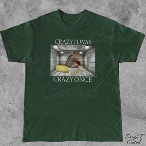 Puede incluir: Camiseta verde oscuro con un gr&aacute;fico de una rata en una habitaci&oacute;n con queso y el texto "CRAZY? I WAS CRAZY ONCE". La rata lleva un guante de boxeo rojo. La camiseta est&aacute; hecha de un material suave.