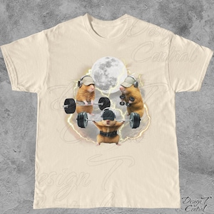 May include: Cream-colored t-shirt featuring a whimsical graphic of three hamsters lifting weights under a full moon. The hamsters wear hats and headphones, with lightning bolts around them. The shirt has a crew neck and short sleeves.