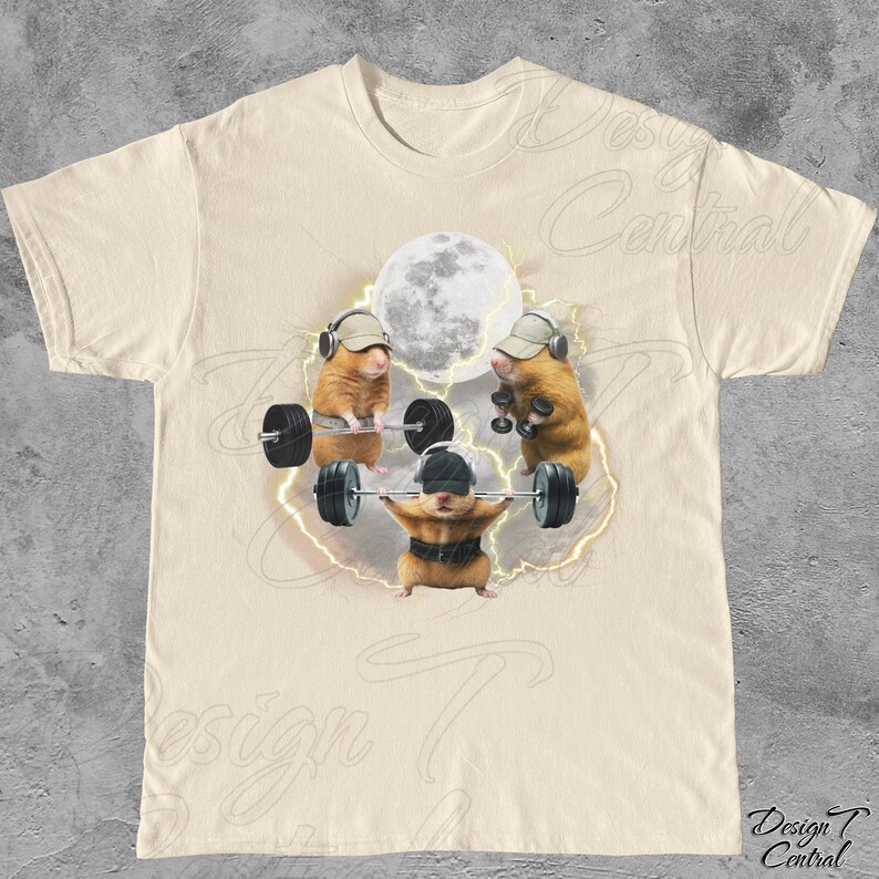 May include: Cream-colored t-shirt featuring a whimsical graphic of three hamsters lifting weights under a full moon. The hamsters wear hats and headphones, with lightning bolts around them. The shirt has a crew neck and short sleeves.