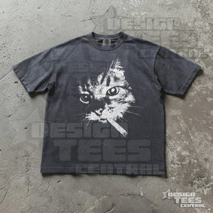 May include: Dark gray t-shirt featuring a white graphic of a cat with a cigarette in its mouth. The cat's face is detailed with whiskers and expressive eyes. The shirt has a vintage, worn look.