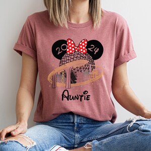 Pu&ograve; includere: Una maglietta color malva con una grafica di una testa di Topolino con un fiocco rosso e il testo "2024" sulle orecchie. La grafica include anche una rappresentazione del giro in Spaceship Earth a Epcot con il testo "Auntie" sotto.