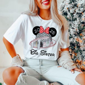 Pu&ograve; includere: Maglietta bianca con un'illustrazione in bianco e nero di una fascia per capelli con orecchie di Topolino con un fiocco rosso e pois bianchi. La fascia &egrave; sopra un'illustrazione in bianco e nero di una sfera con una struttura a strisce rosse e bianche. Il testo "Big Sister" &egrave; sotto l'illustrazione.