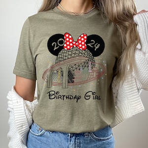 Pu&ograve; includere: Una maglietta verde con un fiocco rosso e bianco a pois, un'illustrazione in bianco e nero di una sfera con una pista intorno e il testo "2024 Birthday Girl".
