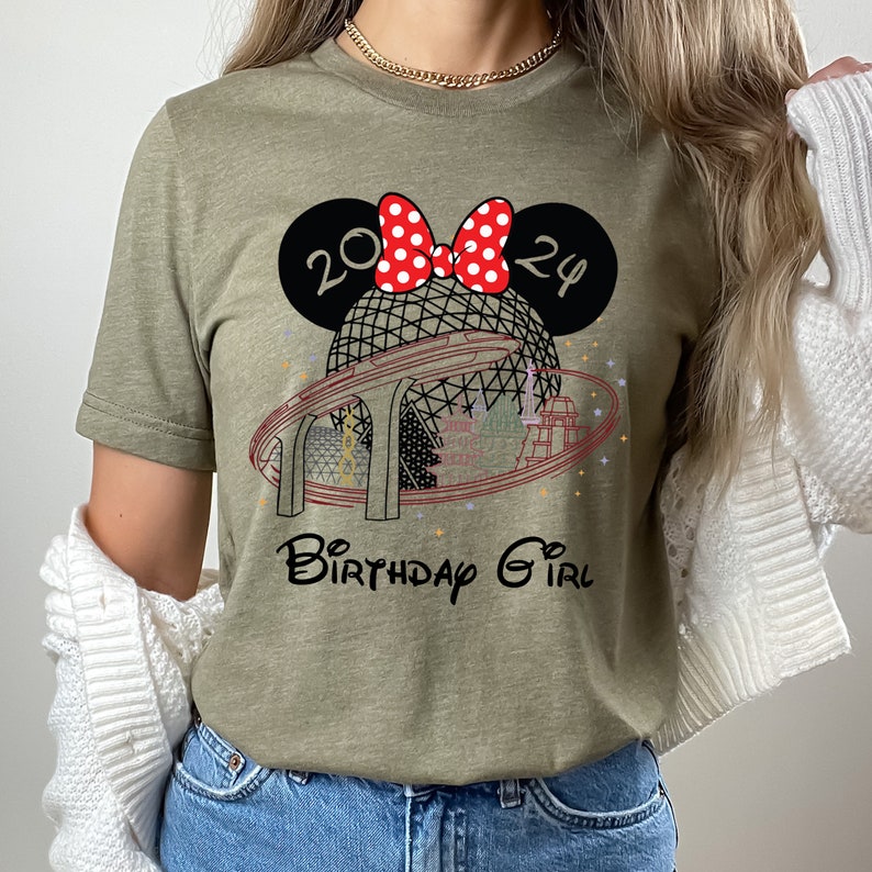 Pu&ograve; includere: Una maglietta verde con un fiocco rosso e bianco a pois, un'illustrazione in bianco e nero di una sfera con una pista intorno e il testo "2024 Birthday Girl".