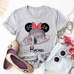 Pu&ograve; includere: Maglietta grigia con un design di orecchie di Minnie Mouse rosse e bianche, una grafica del giro Spaceship Earth a Epcot, e il testo "2024" e "Rachel".