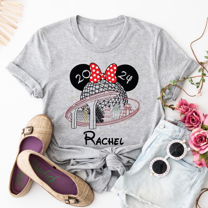 Pu&ograve; includere: Maglietta grigia con un design di orecchie di Minnie Mouse rosse e bianche, una grafica del giro Spaceship Earth a Epcot, e il testo "2024" e "Rachel".