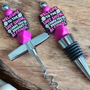 May include: A pink and black bottle stopper with the text "Welcome to the Shit Show, I hope you brought alcohol" and a matching pink and silver corkscrew.