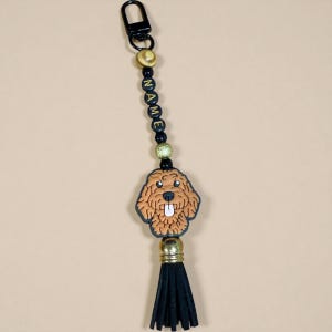 May include: A keychain with a brown dog charm and a black tassel. The keychain has black beads spelling "NAME" and gold-coloured accents. A black clasp is at the top.