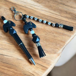 May include: Black and white beaded keychain with a black and white game controller charm and a black tassel. The keychain has white beads with the name "CHARLIE" spelled out on them.