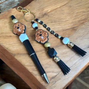 May include: A black pen with a white and gold charm of a brown dog. A gold key chain with a brown dog charm, black and gold beads, and a white bead with a black tassel.