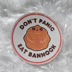 May include: A round sticker with a white background and a red border. The sticker features a cartoon brown character with a happy face and the text "Don't Panic Eat Bannock".