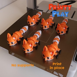 May include: Orange and white articulated fox toys with black feet and eyes are arranged on a brown surface. The text "Printy Play" is in the upper right corner. The text "No supports" and "Print in place" are also visible.