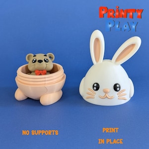 Può includere: Un coniglio pasquale stampato in 3D con un naso rosa e orecchie marroni. Il coniglio è bianco e ha occhi neri. Il coniglio è stampato in posizione, il che significa che non sono necessari supporti. Il coniglio è un guscio vuoto e può essere utilizzato per contenere piccoli oggetti.  Il coniglio è accanto a un uovo rosa stampato in 3D con un orso marrone all'interno. L'orso indossa un papillon rosso. L'uovo è anche stampato in posizione, il che significa che non sono necessari supporti. L'uovo è un guscio vuoto e può essere utilizzato per contenere piccoli oggetti.  Il testo "PRINTY PLAY" è stampato in rosso e bianco su uno sfondo blu.