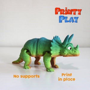Puede incluir: Un juguete de dinosaurio impreso en 3D de color verde, azul y naranja. El dinosaurio es un Triceratops con tres cuernos y una gola. El juguete está articulado y se puede colocar en diferentes posiciones. El texto "PRINTY PLAY" es visible en el fondo. El texto "No supports" y "Print in place" es visible en primer plano.
