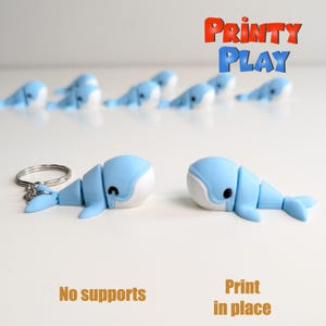 Op de afbeelding: Lichtblauwe walvis sleutelhanger met een witte buik en zwarte ogen. De walvis is gesegmenteerd en heeft een zilveren sleutelring. De afbeelding toont ook verschillende andere walvisbeeldjes en de tekst "Printy Play".