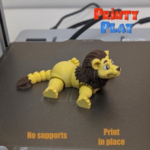 May include: A yellow and brown 3D printed articulated lion figurine. The lion is lying on its belly with its head turned to the side. The text "Print in place" is visible in the image.