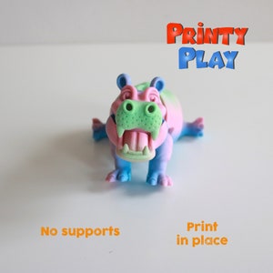 Può includere: Una colorata statuetta di ippopotamo nei toni del blu, rosa e verde. L'ippopotamo ha la bocca aperta con denti visibili ed è etichettato "Printy Play". Sono visibili anche i testi "No supports" e "Print in place".