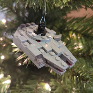 May include: A gray Millennium Falcon ornament made of interlocking toy bricks, hanging from a Christmas tree. The ornament has red and black accents and is suspended by a silver wire.