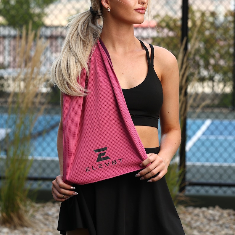 May include: A pink microfiber towel with the word "ELEV8T" and a stylized letter "E" printed in black. The towel is being held by a person wearing a black sports bra and a black skirt.