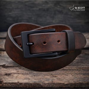 May include: A brown leather belt with a black buckle. The belt is made of a textured, dark brown leather and has a rectangular black buckle. The belt is coiled on a wooden surface. The text "B. SCOTT LEATHER" is in the upper right corner.