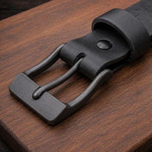 May include: A black leather belt featuring a matte dark gray metal buckle and a matching leather keeper loop. The belt strap has a smooth texture and is secured by a single prong that passes through a circular hole in the leather material.