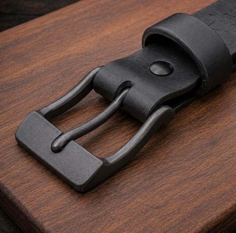 May include: A black leather belt featuring a matte dark gray metal buckle and a matching leather keeper loop. The belt strap has a smooth texture and is secured by a single prong that passes through a circular hole in the leather material.