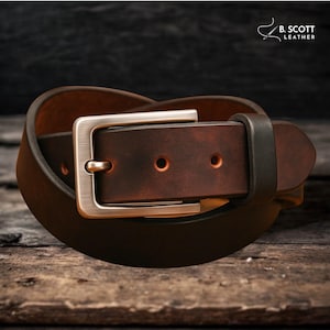 May include: A rolled brown leather belt with a silver-colored buckle. The belt has two small holes and is displayed on a wooden surface. The brand name "B. Scott Leather" is visible in the upper right corner.