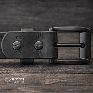 May include: A black leather belt featuring a matte black metal buckle and two star-shaped screw fasteners securing the strap. The leather has a visible grain texture, and the brand name B. SCOTT LEATHER is printed in white in the corner.