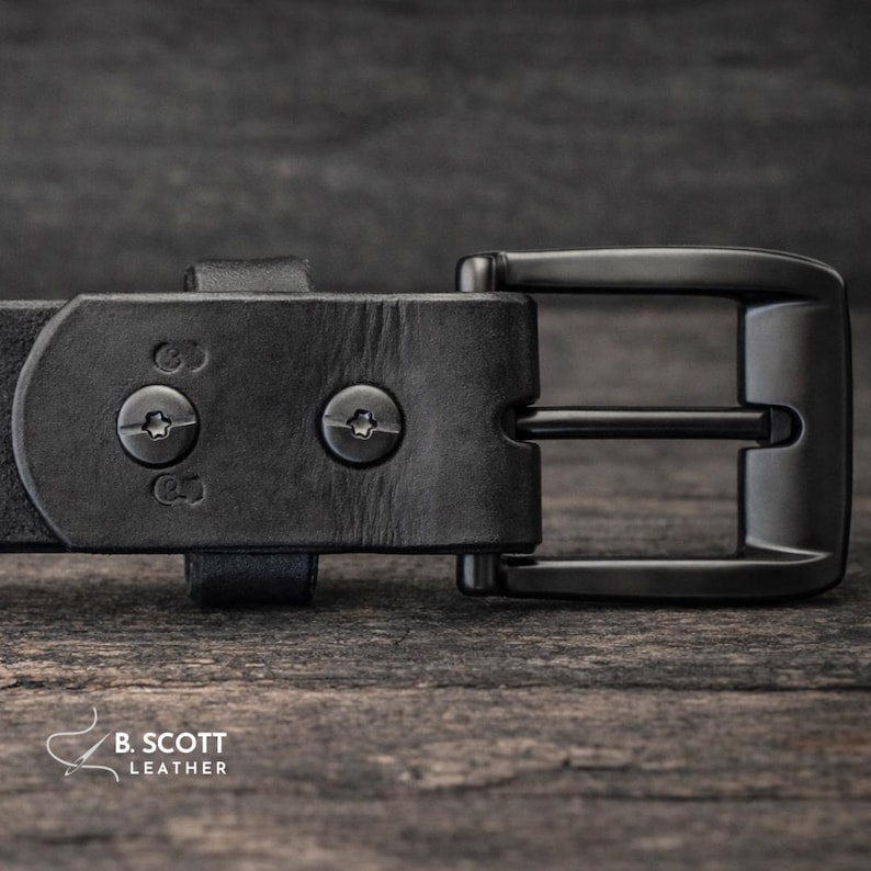 May include: A black leather belt featuring a matte black metal buckle and two star-shaped screw fasteners securing the strap. The leather has a visible grain texture, and the brand name B. SCOTT LEATHER is printed in white in the corner.