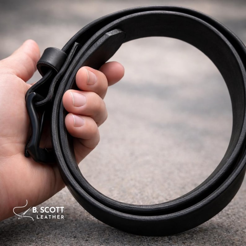 May include: A black leather belt featuring a matte black buckle and a matching leather keeper loop. The belt is coiled into a circular shape. The text B. SCOTT LEATHER is printed in white in the bottom left corner of the image.